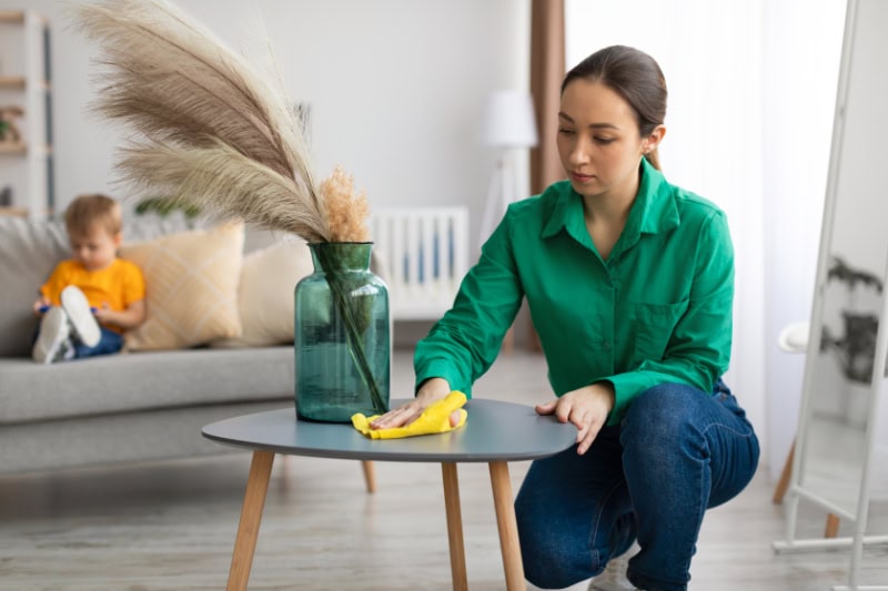 Spring cleaning. Young mom dusting off coffee table and tidying up in living room while her toddler son watching cartoon on cellphone, sitting on sofa on background.